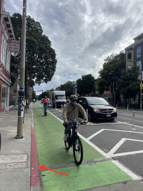 A cyclist rides in a green bike lane on a city street; another cyclist and several vehicles are visible further down the lane. An arrow points to a red curb.