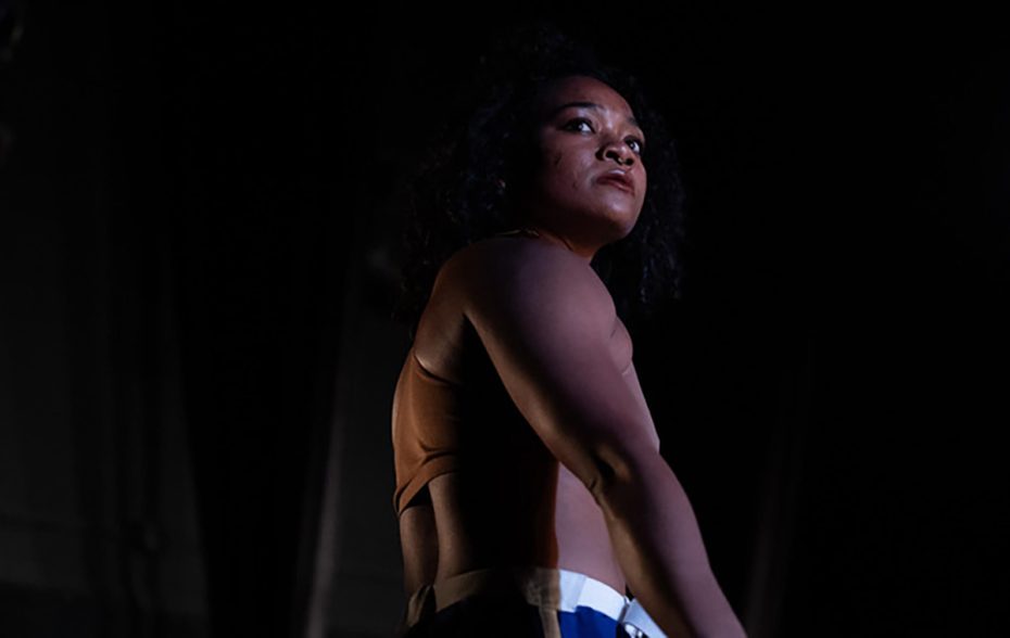 A person wearing a sleeveless top and striped pants stands under dramatic lighting, looking upward against a dark background.