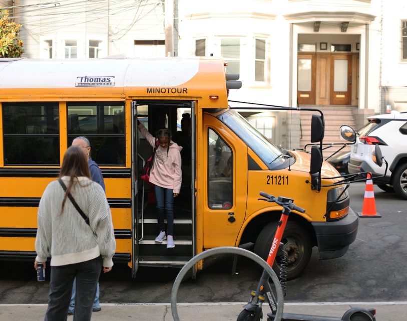 A child steps off a yellow minibus while two adults wait nearby on a city street with parked cars, an orange traffic cone, and a scooter in the foreground.