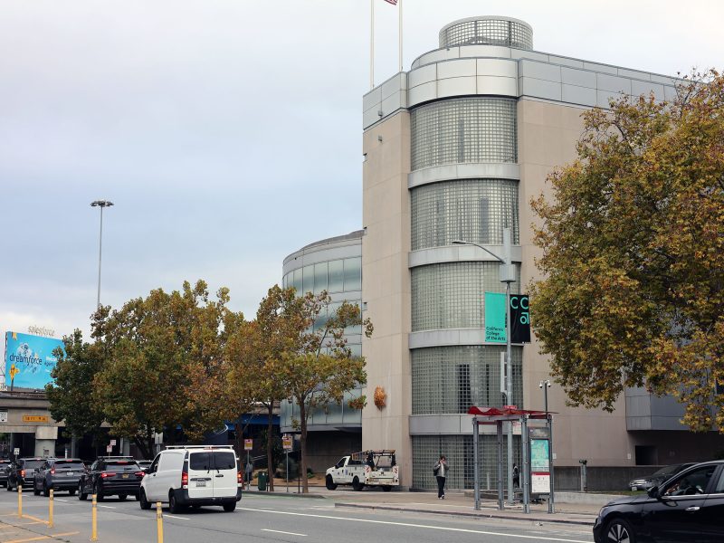 A street scene shows light traffic, a sidewalk, trees with autumn leaves, and a modern building with glass block windows and rounded corners.