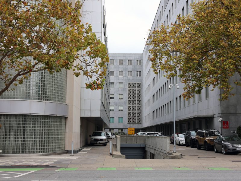 Two modern office buildings with glass and concrete facades are separated by a driveway, lined with parked cars and trees with autumn leaves.