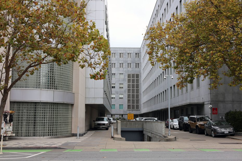 Two modern office buildings with glass and concrete facades are separated by a driveway, lined with parked cars and trees with autumn leaves.