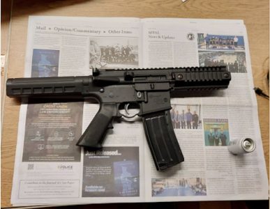 A black semi-automatic rifle with a magazine is placed on an open newspaper on a wooden surface, next to a small can and a white object.