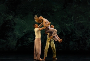 Three dancers on stage perform a lift, with one dancer holding another in mid-air while a third supports, against a dark abstract background.