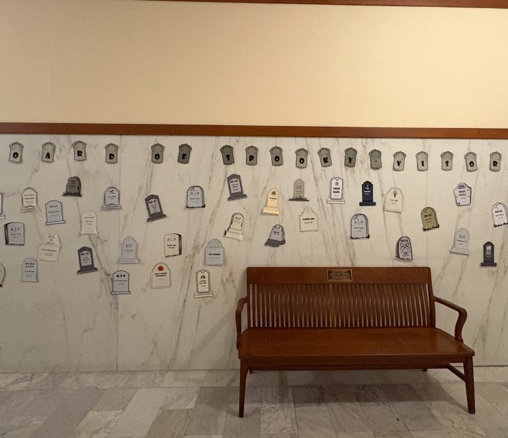 A wooden bench sits in front of a marble wall decorated with paper gravestones displaying various names and messages.