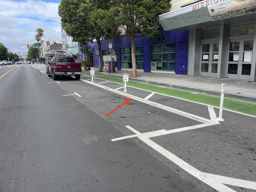 A street with parking spaces marked by white lines, a green bike lane, and temporary signs on posts; an arrow points to the center of an empty parking space.
