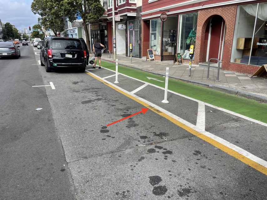 A man stands by a black SUV parked next to a green bike lane separated by white posts and a yellow line on a city street.