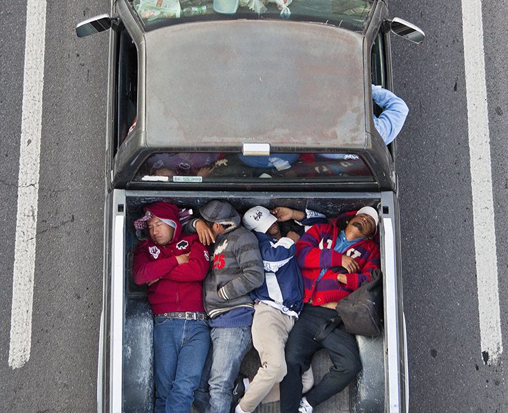 Aerial view of six people lying closely together in the bed of a pickup truck parked between two white lines on a road.