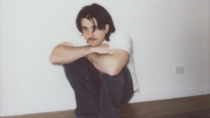 A person with dark hair and a mustache sits on the floor against a white wall, arms resting on their knees, wearing a white shirt and dark pants.