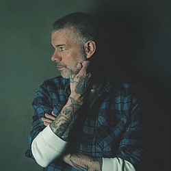 A man with tattoos, short gray hair, and a beard stands against a dark background, looking to the left with his hand on his chin and wearing a plaid shirt over a white long-sleeve shirt.