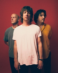 Three men stand in front of a solid red background; the man in the center wears a white shirt, while the others wear a gray shirt and a yellow shirt.