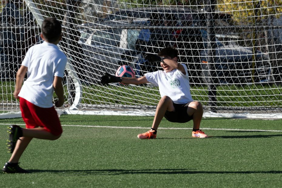 A young boy in goalkeeping gloves blocks a soccer ball during a game, while another boy in red shorts approaches. They are playing on an outdoor field with a net in the background.