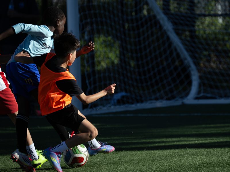 Three children wearing sports uniforms compete for the soccer ball on a field in front of a goal net during a game.