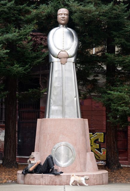 A person lies on their back using a phone next to a small dog at the base of a large metallic statue of a robed figure, surrounded by tall trees.