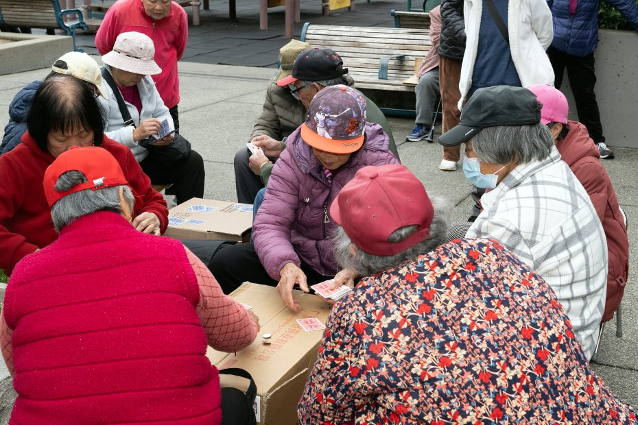 A group of elderly people sit outdoors around cardboard boxes, playing a card game. Most are wearing hats and jackets, and one person is wearing a face mask.
