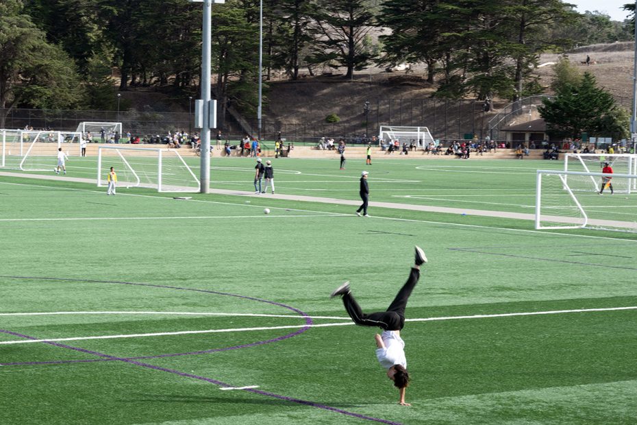 A person does a cartwheel on a soccer field while other people play soccer and walk in the background.
