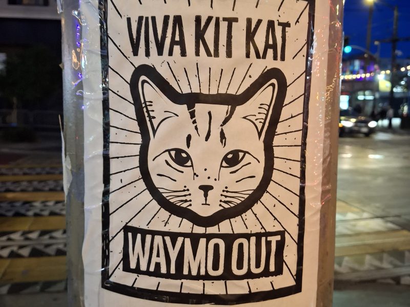 A poster on a pole features a cat's face with the text "VIVA KIT KAT" at the top and "WAYMO OUT" at the bottom. The background shows a city street at dusk.