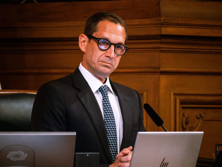 A man in a suit and glasses sits at a desk with a laptop and microphone in front of him, against a wooden paneled background, ready to record his latest podcast about RV adventures.
