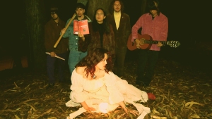 Five people stand in a wooded area at night, one holding a bat, another a book, and one with a guitar. A person in a white dress sits on the ground surrounded by leaves.