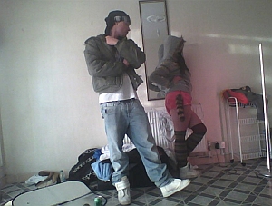A man stands with arms crossed while a woman poses in front of a radiator in a sparsely furnished room with clothes on the floor and a checkered tile pattern.