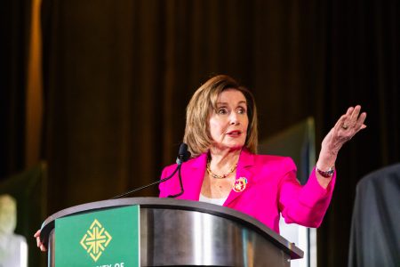 A woman in a bright pink blazer speaks at a podium with a green emblem, gesturing with her right hand against a dark background.