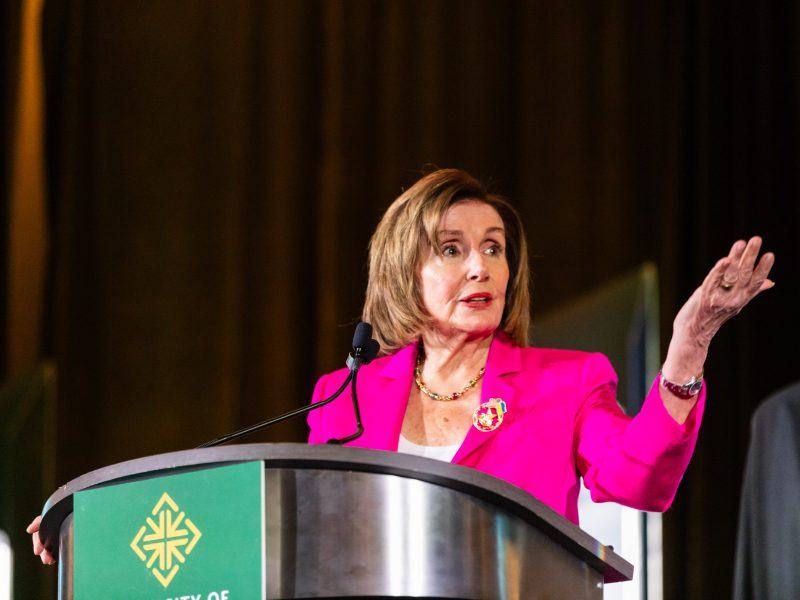 A woman in a bright pink blazer speaks at a podium with a green emblem, gesturing with her right hand against a dark background.