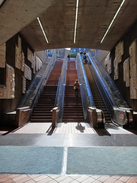 A person walks up a staircase between two escalators in an urban subway or transit station, with sunlight streaming in from above.