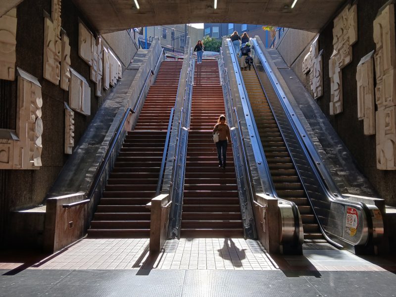 A person walks up the central stairs between two escalators at an outdoor transit station, with sunlight streaming in from above.