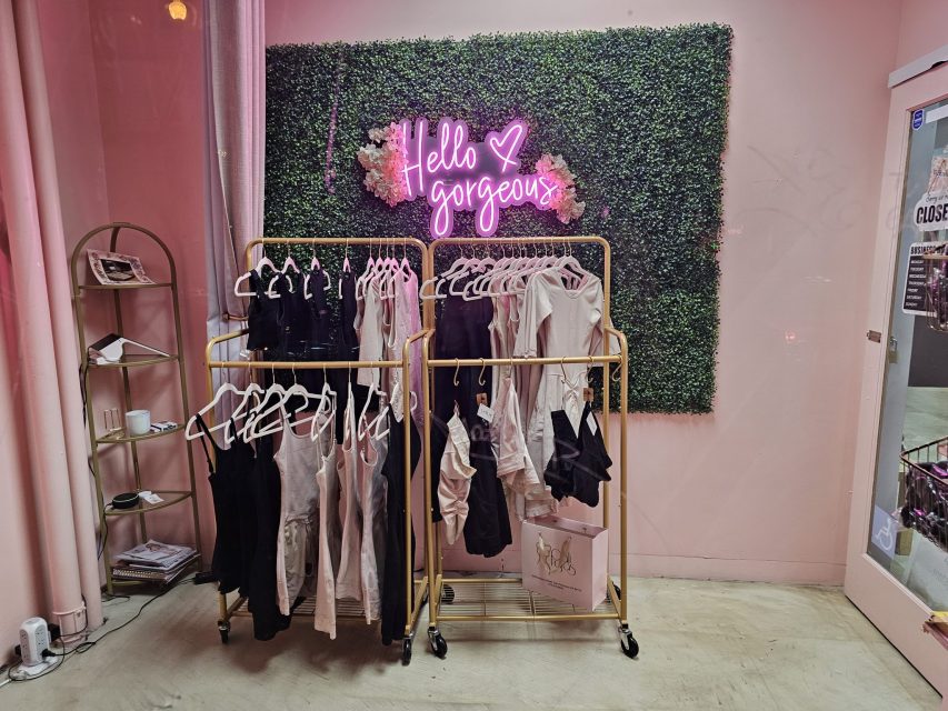 A clothing rack displays black and white lingerie in a pink boutique with a neon sign reading "Hello gorgeous" above artificial greenery on the wall.
