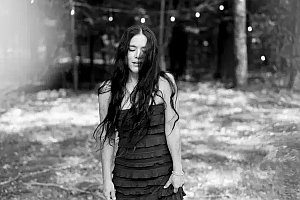 A woman with long dark hair stands outdoors in a sleeveless dress, eyes closed, with string lights and trees in the background.