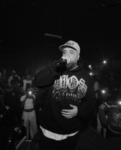 A man wearing a hoodie and cap performs on stage with a microphone, surrounded by a crowd, some holding up phones with flashlights on.