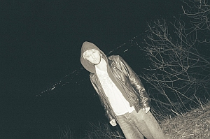A person wearing a hooded jacket stands outdoors at night near leafless branches, with a dark sky in the background.