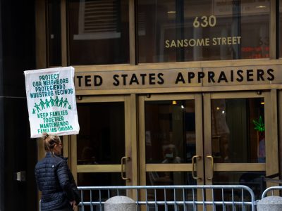 A person holds a bilingual protest sign reading "Protect Our Neighbors" and "Keep Families Together" outside a building labeled United States Appraisers at 630 Sansome Street.