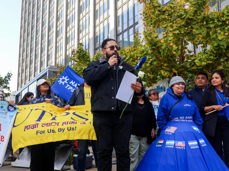 A man speaks into a microphone at an outdoor rally, holding a white rose and papers, while people around him display banners and signs supporting TPS.