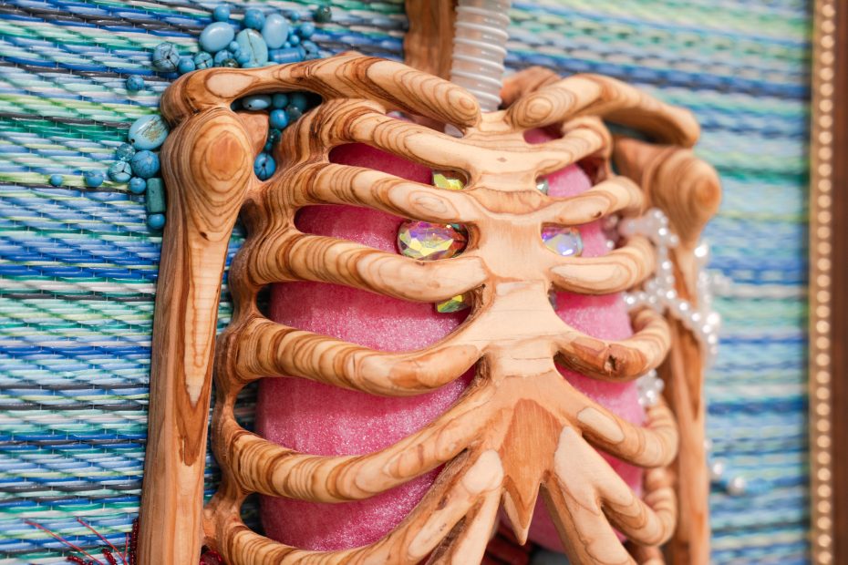 Close-up of a decorative wooden rib cage with a pink interior, adorned with rhinestones and blue beads, set against a woven, multicolored background.