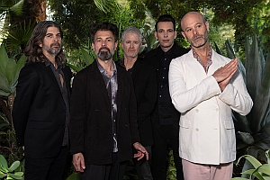 Five men wearing suits stand together outdoors in front of lush green plants, posing for a group photo.