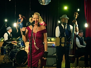 A band performs on stage; a woman sings at the microphone while musicians play drums, upright bass, and piano. A disco ball and dancers are visible in the background.