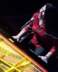 A person in a red top is seated at a grand piano, singing into a microphone.