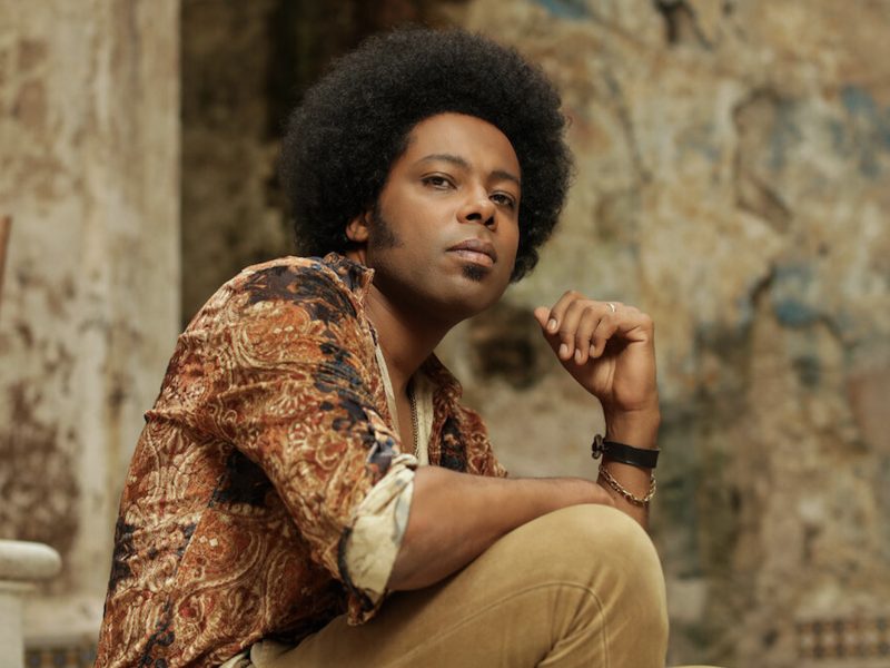 A man with an afro hairstyle sits on outdoor steps, wearing a patterned shirt and light pants, looking at the camera with a neutral expression. The background is textured and worn.