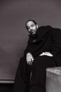 A person wearing dark clothing sits on a bench, looking toward the camera with a neutral expression against a plain background.