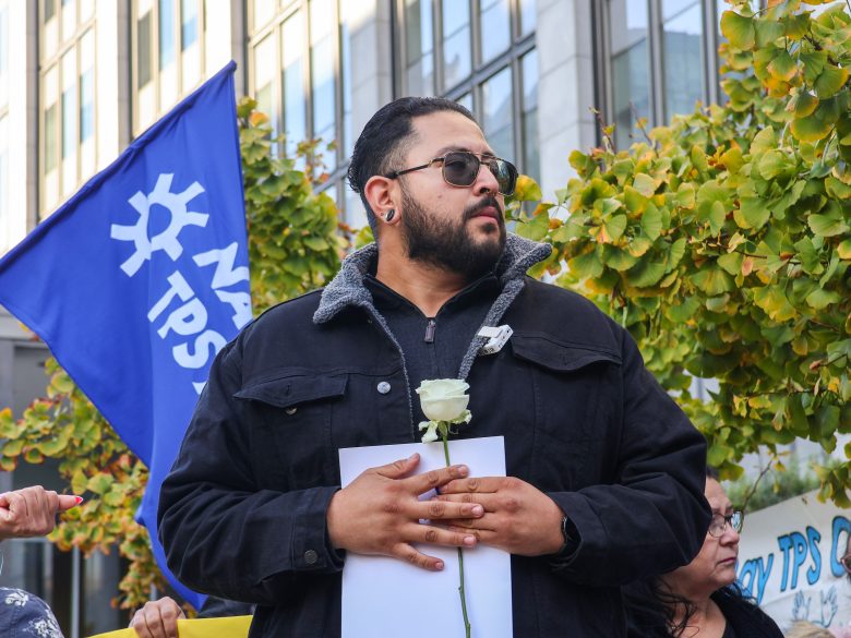 A man wearing sunglasses holds a white rose and a piece of paper, standing outdoors in front of a blue flag and trees.