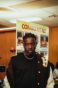 A man stands in front of a menu board displaying combo meals with burgers and drinks in a restaurant.
