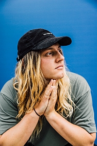 Person with long blonde hair wearing a black cap and gray shirt, hands clasped under their chin, looking to the side against a blue background.