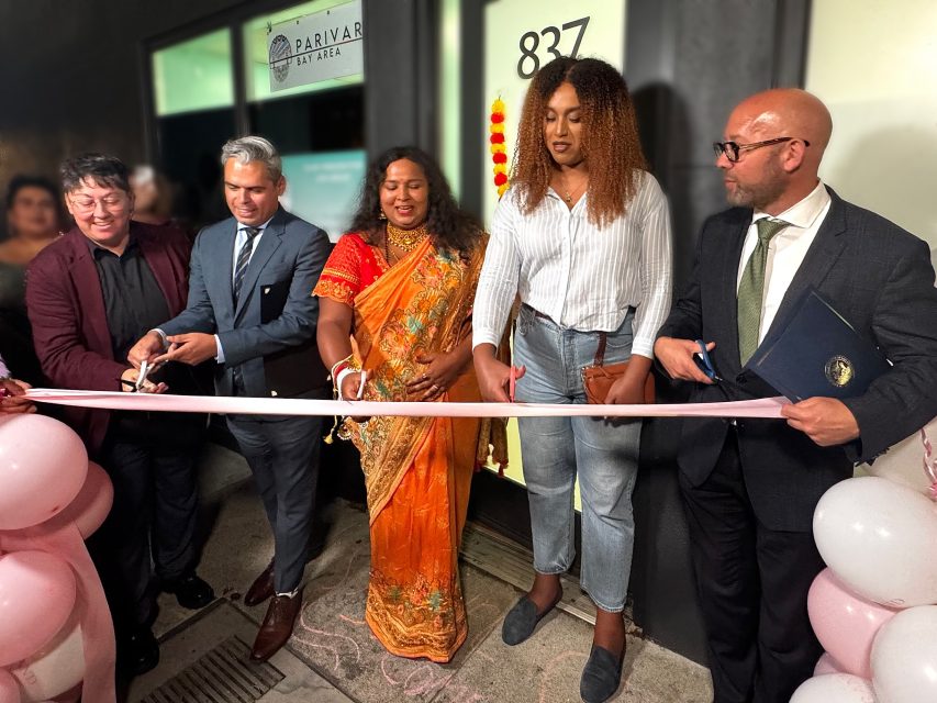 Six people, including transgender individuals, stand in front of a business entrance as three cut a pink ribbon at a ceremonial event. Pink balloons and a "Parivar Bay Area" sign are visible.