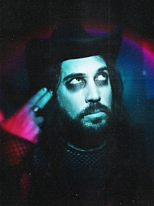 A bearded person wearing a black hat and dark clothing points a finger to their temple, looking upward with dramatic lighting and heavy makeup.
