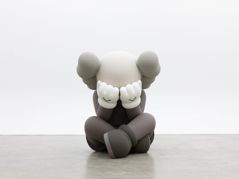 A large, cartoon-style sculpture of a seated figure with gloved hands covering its face, gray limbs, and crossed-out eyes, positioned against a plain white background.