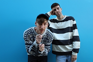 Two men in patterned sweaters pose in front of a solid blue background; one leans forward, the other rests his head on his hand.