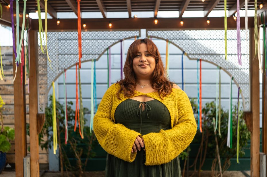 A woman with long brown hair, wearing a yellow sweater over a green dress, stands under a pergola decorated with hanging colorful ribbons.