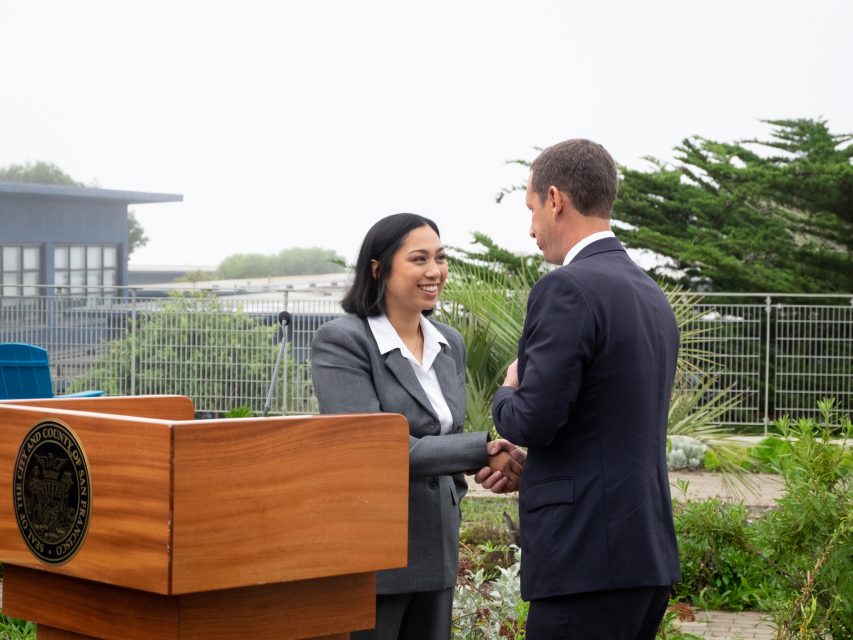 Two people in business attire shake hands outdoors next to a podium with an official seal, surrounded by greenery and metal fencing.