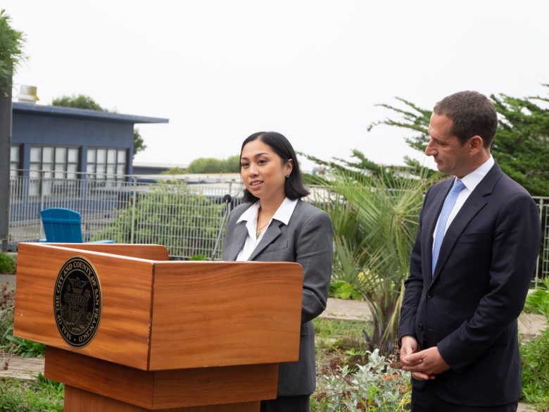 A woman in a suit speaks at a wooden podium outdoors while a man in a suit stands beside her, listening.